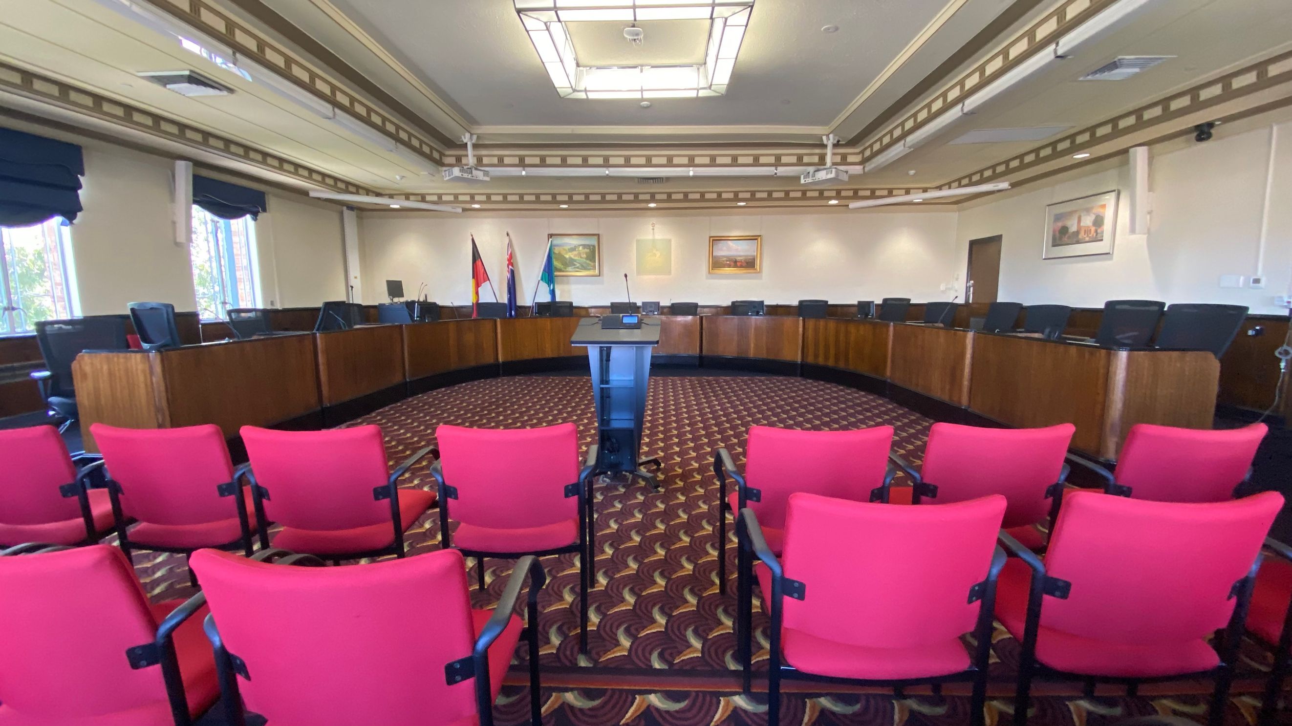 A conference room with a patterned carpet and a large U-shaped wooden table at the back, surrounded by black chairs. In the centre, there's a podium with a microphone. Red chairs are arranged in rows facing the podium. The walls have framed paintings and flags are displayed behind the table. The ceiling features a large rectangular light fixture, and windows with dark curtains are on the left.