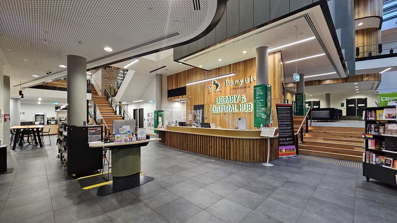 On the left, there is a black wall with a "Fire Indicator Panel" sign and a door handle. In the middle, glass entrance doors with green stripes and a small black mat on the floor, leading to a reception area with wooden counters and bookshelves. On the right, a colourful recycling station with labelled bins and a wall with various posters. The floor is covered with large grey tiles, and the ceiling is white with recessed lighting and perforated panels.