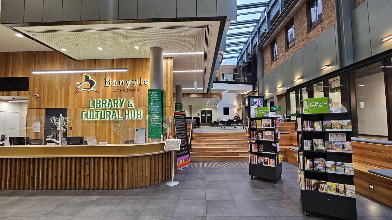 On the left, there is a wooden reception counter with the text "Banyule City Council Library & Cultural Hub" on the wall behind it, along with a green information board. In the middle, a set of wooden steps leads to an open seating area with tables and chairs. On the right, there are bookshelves filled with books and a green sign. The floor is covered with large grey tiles, and the ceiling is white with recessed lighting and a skylight above the seating area.