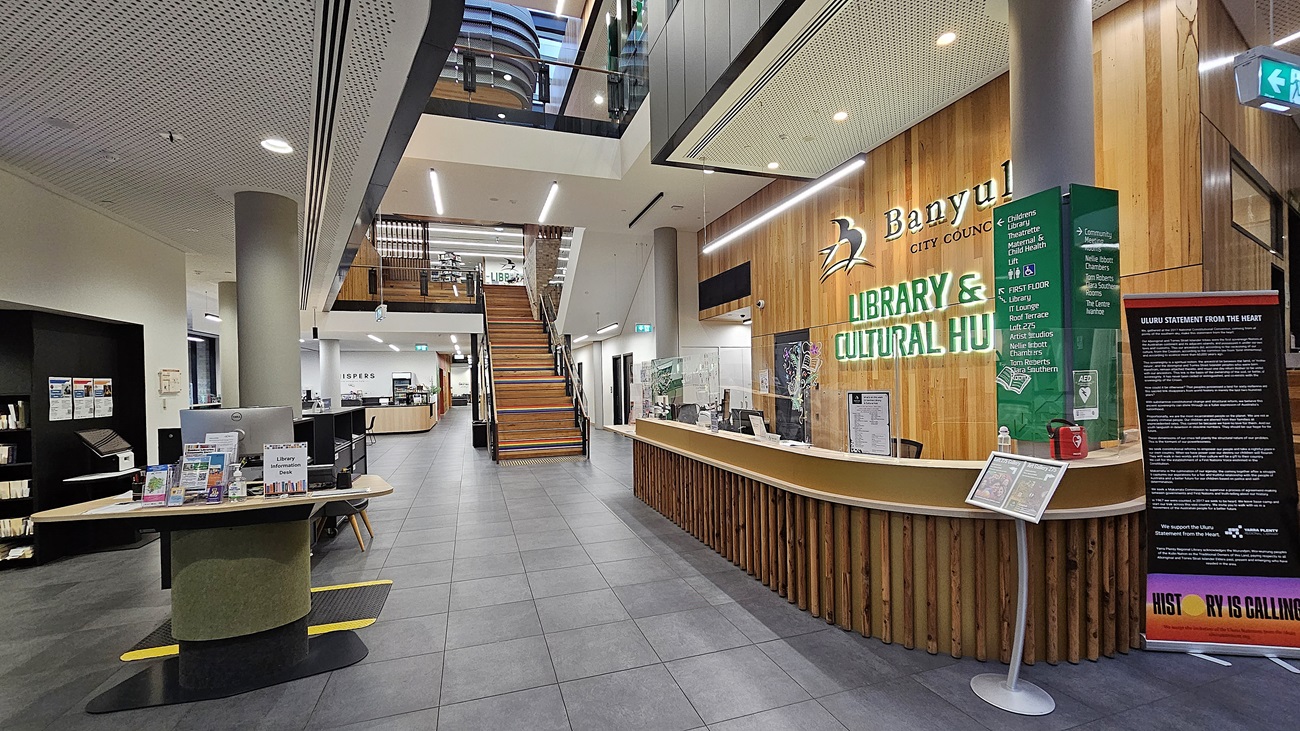 On the left, there is a small table with brochures and a cushioned seat, next to a black wall with bookshelves. In the middle, a staircase with wooden steps leads to the upper floor, with a glass railing and a skylight above. On the right, a long wooden reception counter with the text "Banyule City Council Library & Cultural Hub" on the wall behind it, along with green information boards. The floor is covered with grey tiles, and the ceiling is white with perforated panels and recessed lighting.
