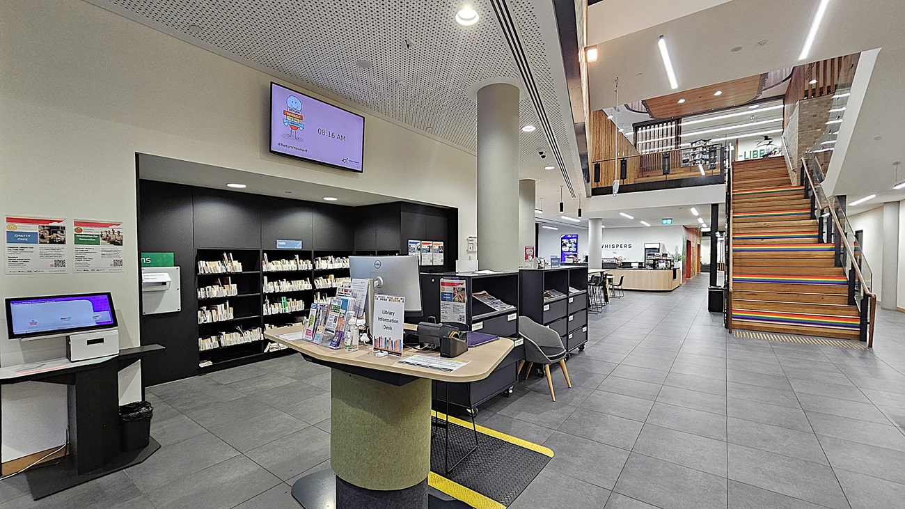 On the left, there is a self-service kiosk with a touchscreen and a small table with brochures. Next to it, black bookshelves filled with books. In the middle, a round information desk with a computer and more brochures. On the right, a wide staircase with wooden steps and a glass railing leading to the upper floor. The background includes more bookshelves and a reception counter. The floor is covered with large grey tiles, and the ceiling is white with perforated panels and recessed lighting.