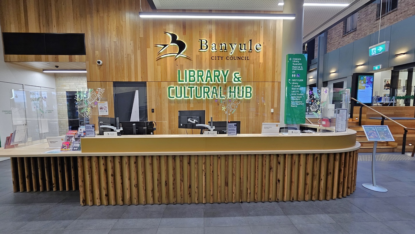 On the left, there is a white wall with a small window and a black shelf. In the middle, a curved wooden reception counter with clear protective screens and various brochures. Behind the counter, a wooden wall with the text "Banyule City Council Library & Cultural Hub" and a logo. On the right, a staircase with wooden steps and a glass railing leading to the upper floor. The floor is covered with large grey tiles, and the ceiling is white with recessed lighting and perforated panels.
