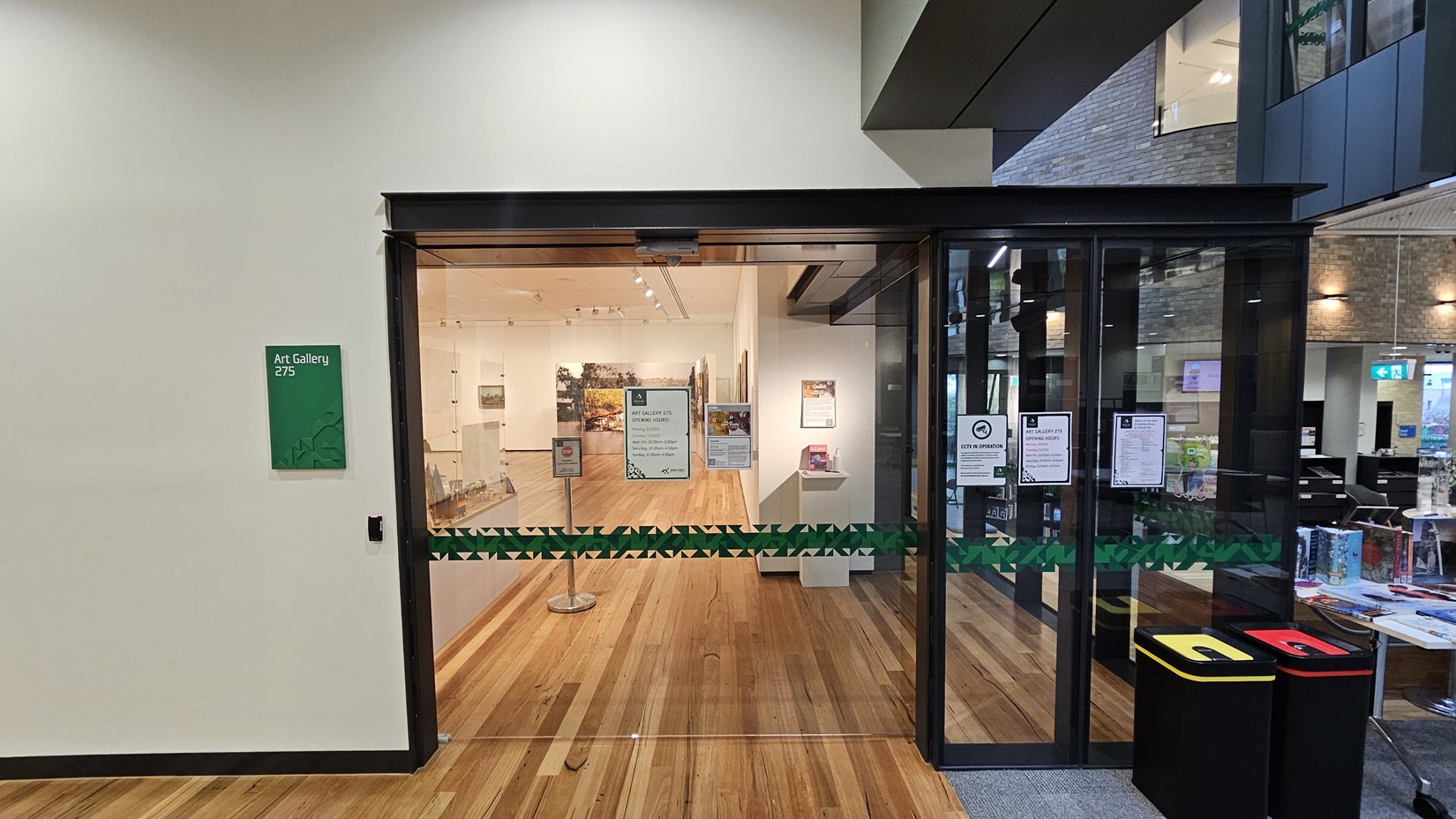 On the left, there is a green sign reading "Art Gallery 275" next to a glass entrance with a green ribbon across it. In the middle, there is a wooden floor leading into the gallery, with artworks displayed on the walls inside. In the background, there is a seating area with chairs and a table holding brochures. On the right, there is a wooden wall with a digital screen and a grey carpeted floor. The ceiling is white with recessed lighting and grey panels.