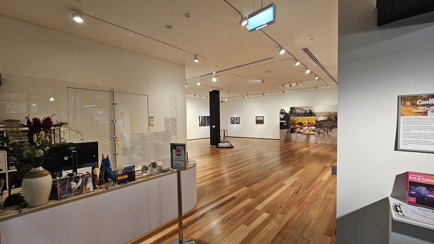 On the left, there is a white reception desk with a clear partition, flowers in a vase, and various brochures and items. In the middle, there is an open space with wooden flooring, leading to an exhibition area with artworks on the walls. In the background, there are more artworks and a display stand. On the right, there is a white wall with a framed information board and a small shelf holding brochures. The ceiling is white with recessed lighting and track lights.