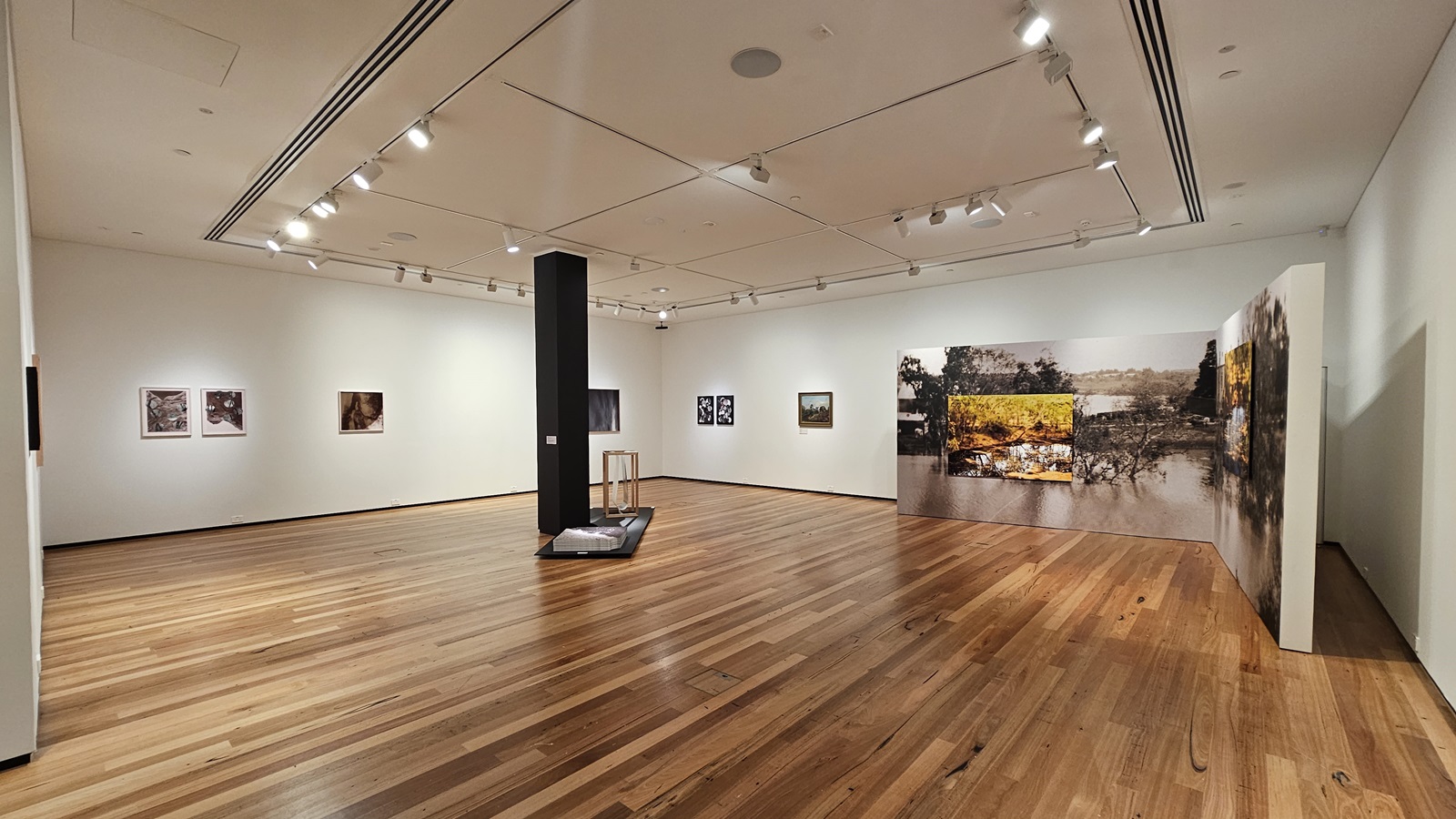 On the left, there are several framed artworks hung on a white wall. In the middle, there is a black column with a small display stand next to it, and a low platform with an object on it. In the background, more framed artworks are displayed on the walls. On the right, there is a large mural depicting a landscape scene. The floor is made of polished wooden planks, and the ceiling is white with recessed lighting and track lights.