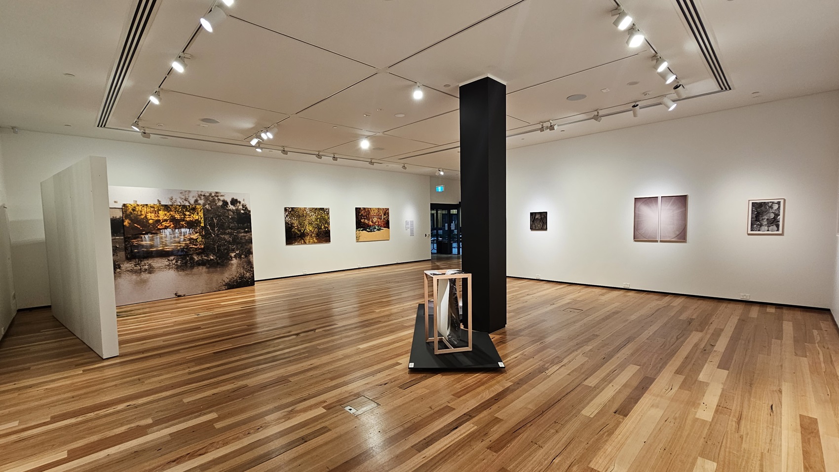 On the left, there are large framed photographs hung on a white wall. In the middle, there is a black column with a small display stand featuring a geometric sculpture. In the background, more framed photographs are displayed on the walls. On the right, there are two framed photographs on a white wall. The floor is made of polished wooden planks, and the ceiling is white with recessed lighting and track lights.