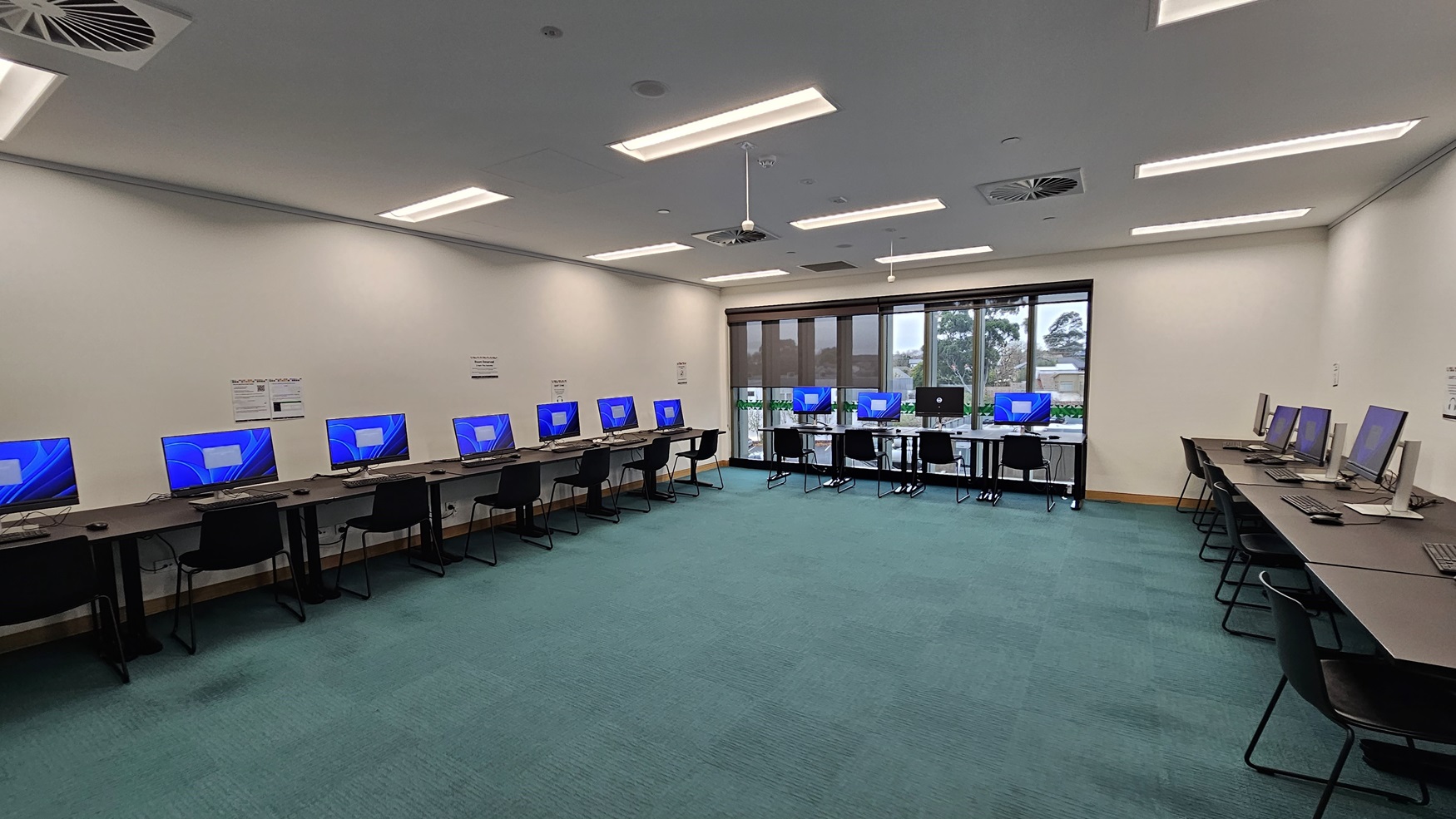 On the left, there are two self-service kiosks on black stands, a green "Library" sign with a bird logo, and two bins, one black and one yellow. In the middle, there is a glass double door leading to a room with green carpet and black chairs. On the right, there are bookshelves filled with books and magazines. The floor is covered in grey carpet tiles, and the ceiling has white perforated panels with recessed lighting and black linear lights.