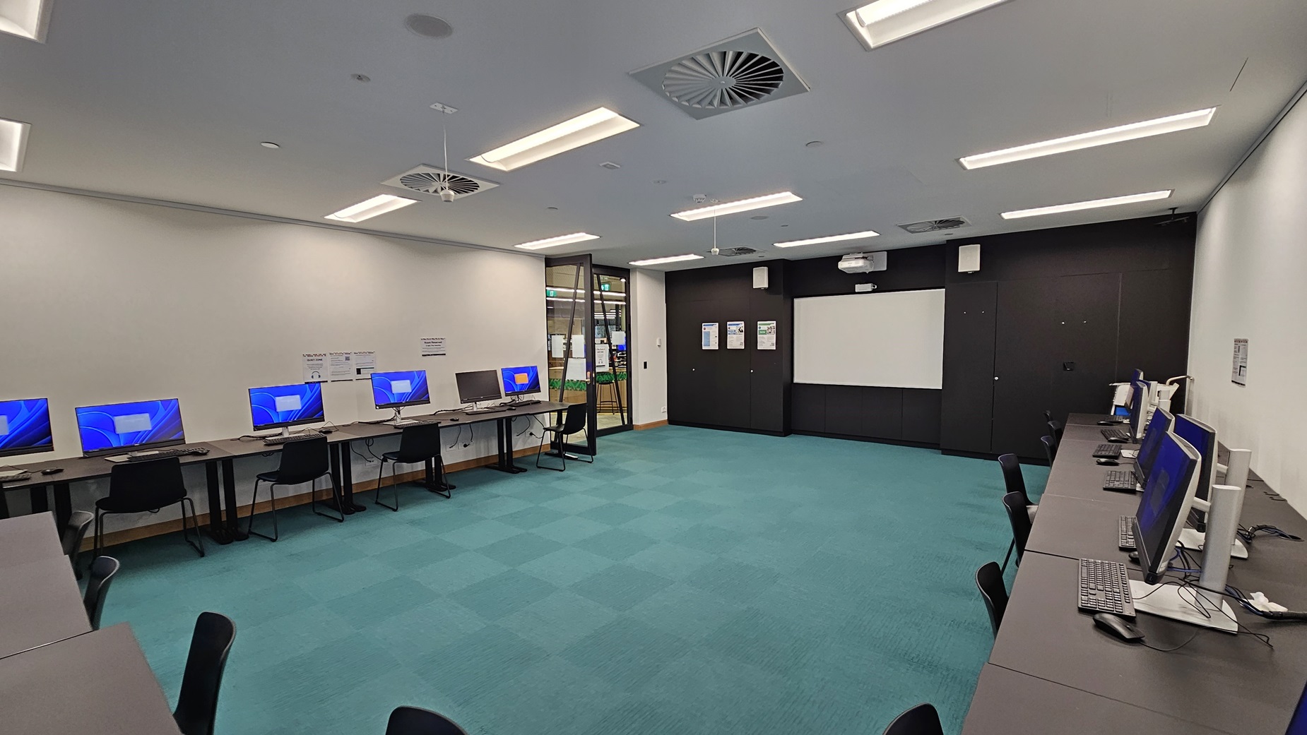 On the left, there are several computer workstations with blue screens and black chairs lined up against a white wall. In the middle, there is an open space with a turquoise carpet. In the background, there is a glass door leading to another room, flanked by black walls with notices and a whiteboard. On the right, there are more computer workstations with blue screens and black chairs. The ceiling is white with recessed lighting and ventilation units.