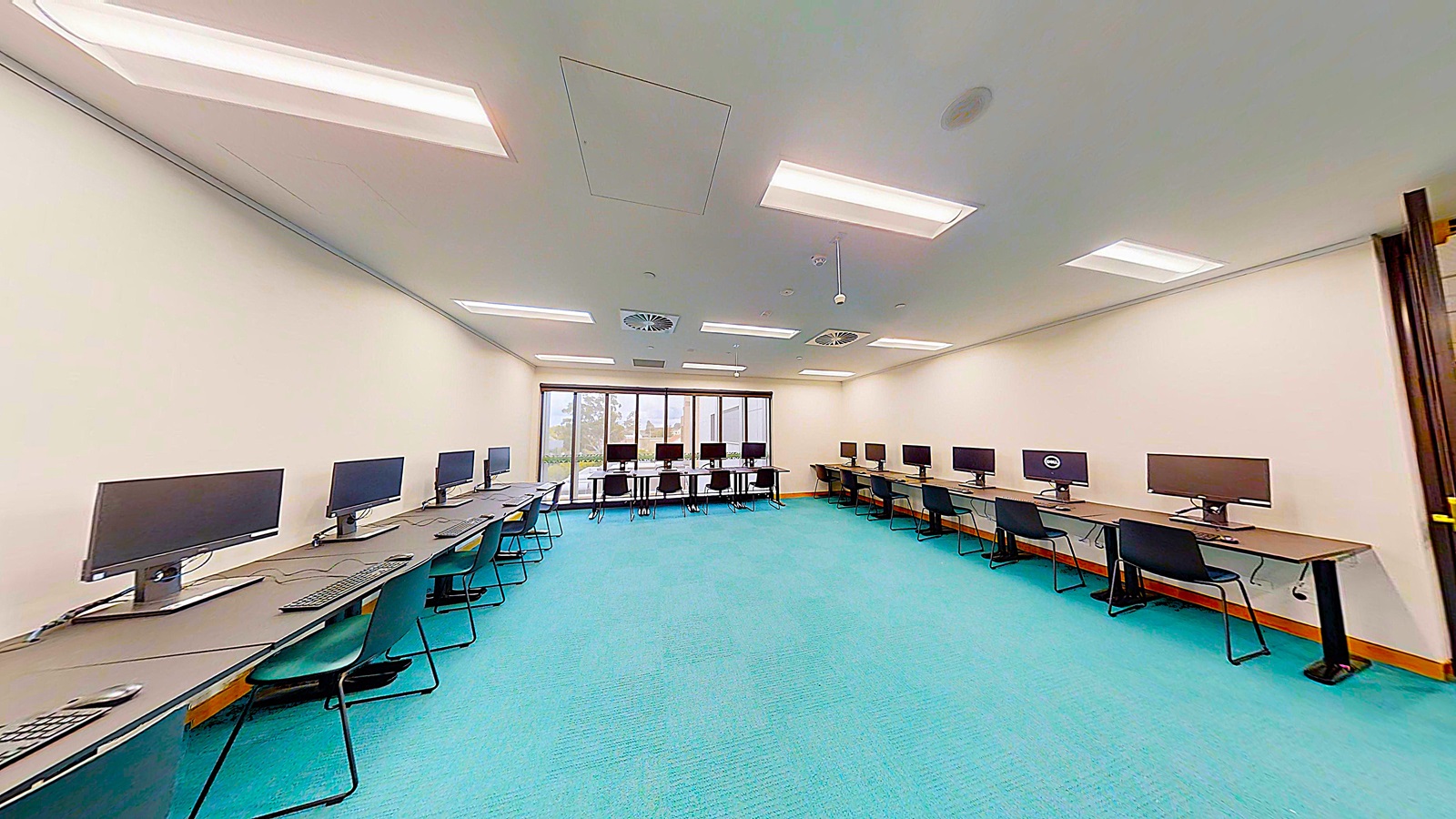 On the left, there are several computer workstations with black chairs and monitors arranged along a long desk against a white wall. In the middle, there is an open space with a turquoise carpet. On the right, there are additional computer workstations with black chairs and monitors along another long desk against a white wall. The ceiling is white with recessed lighting and ventilation units.