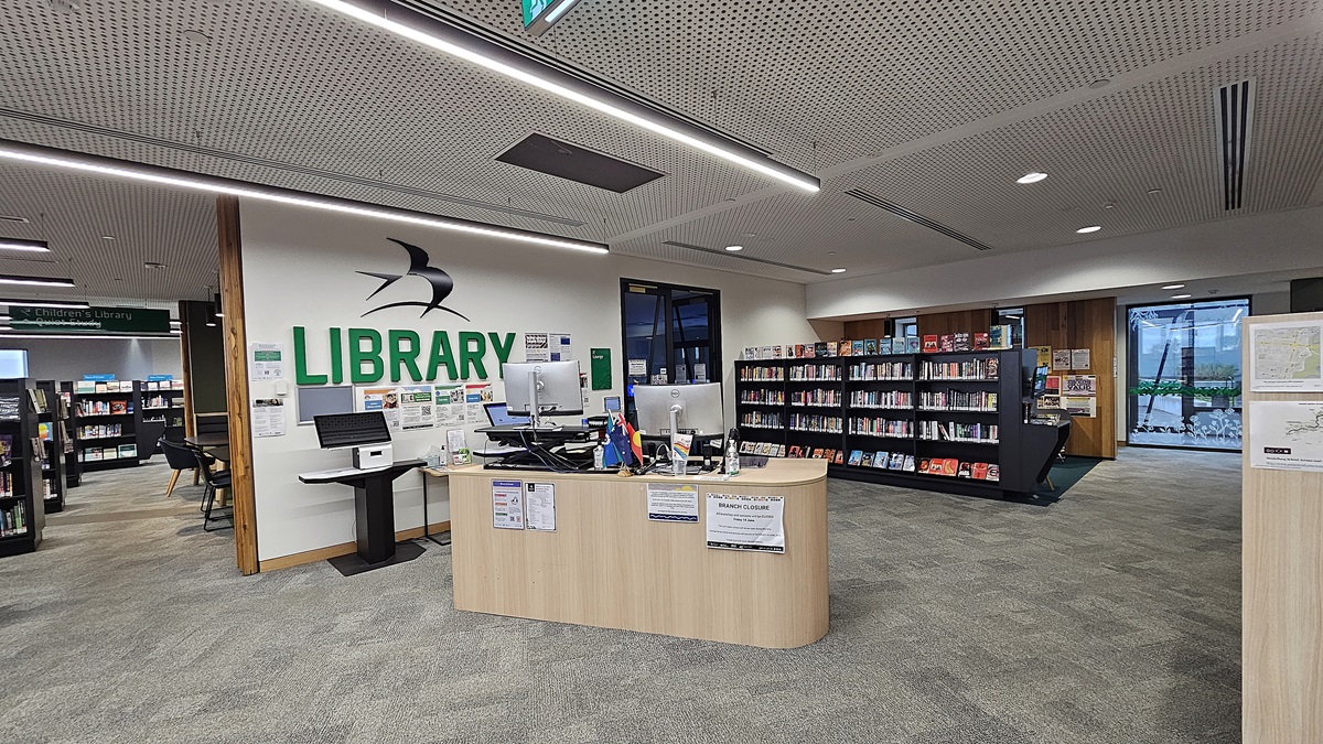 On the left, there is a white wall with the word "LIBRARY" in green and a black and white bird logo above a computer desk with monitors and office supplies. In the middle, there is a curved wooden reception desk with notices and a computer. On the right, there are bookshelves filled with books and a large window with a view of trees. The floor is covered in grey carpet tiles, and the ceiling is white with perforated panels and recessed lighting.