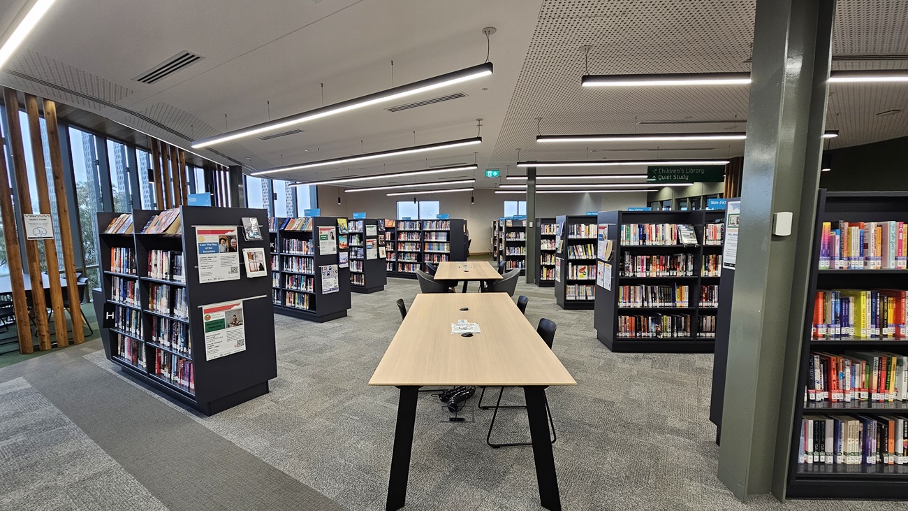On the left, there are black bookshelves filled with books and a wall with vertical wooden slats and large windows. In the middle, there are long wooden tables with black chairs and more bookshelves behind them. On the right, there are additional black bookshelves filled with books and a grey column. The floor is covered in grey carpet tiles, and the ceiling is white with perforated panels and recessed linear lighting.