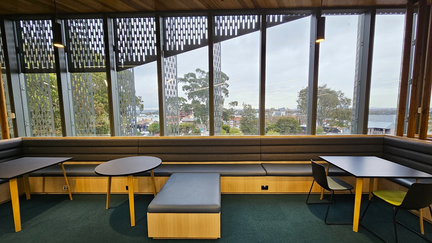 On the left, there is a round table with a yellow base and a grey cushioned bench along the wall. In the middle, there is a long grey cushioned bench with a wooden base and large windows offering a view of trees and buildings. On the right, there are two square tables with black tops, yellow and black chairs, and another section of the grey cushioned bench. The floor is covered in dark green carpet, and the ceiling has wooden panels with recessed lighting.