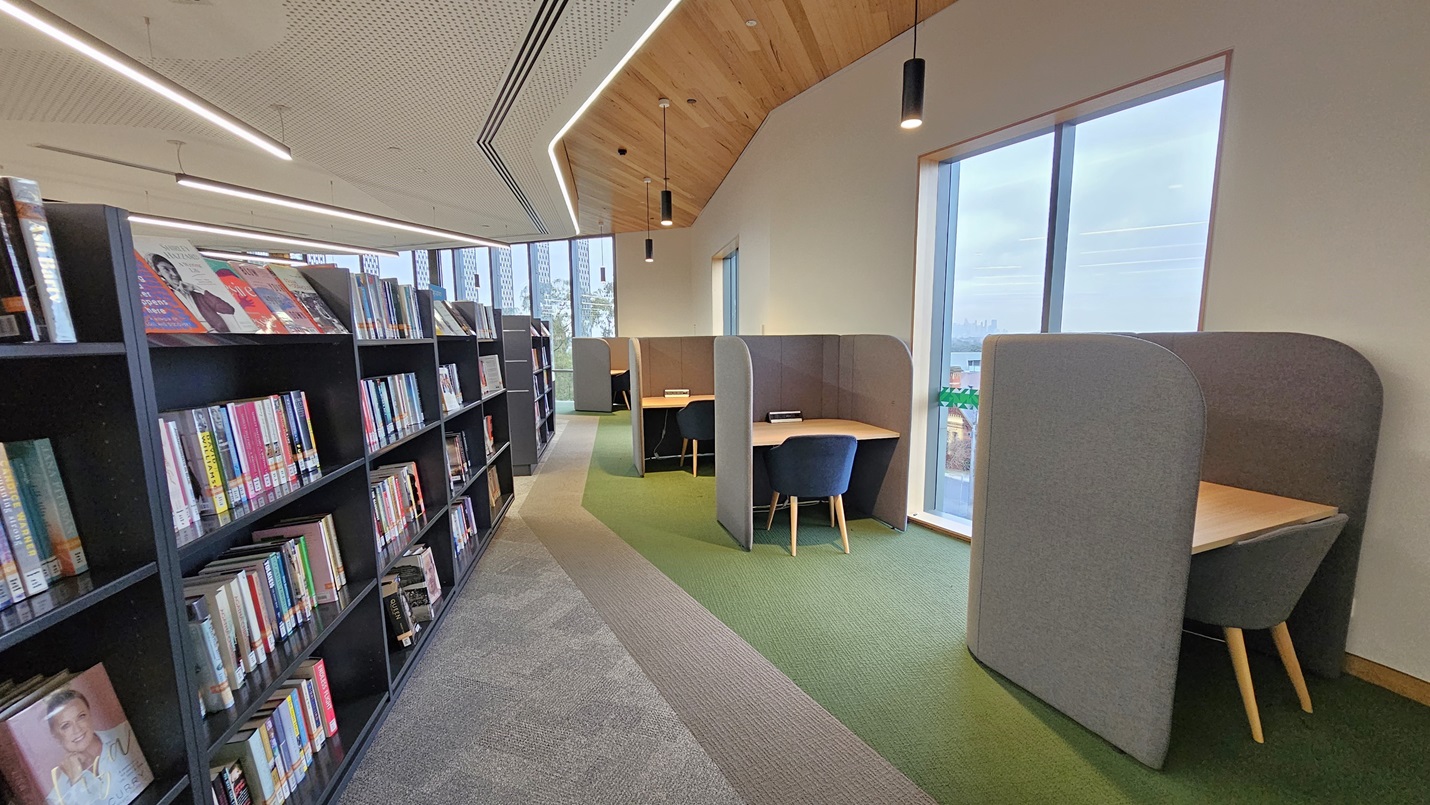 On the left, there are black bookshelves filled with books and magazines. In the middle, there is a walkway with grey and green carpet tiles leading to study cubicles. On the right, there are grey study cubicles with wooden desks and black chairs, positioned next to large windows with a view of buildings. The floor is a mix of grey and green carpet tiles, and the ceiling has white perforated panels and wooden sections with recessed lighting.