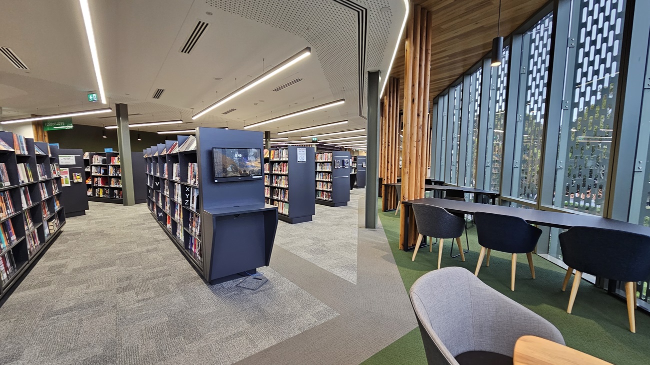 On the left, there are rows of black bookshelves filled with books and a television screen mounted on one of the shelves. In the middle, there is a walkway with grey and green carpet tiles leading to more bookshelves. On the right, there are large windows with vertical wooden slats and a view of trees, along with a row of tables and black chairs. The floor is a mix of grey and green carpet tiles, and the ceiling has white perforated panels with recessed lighting.