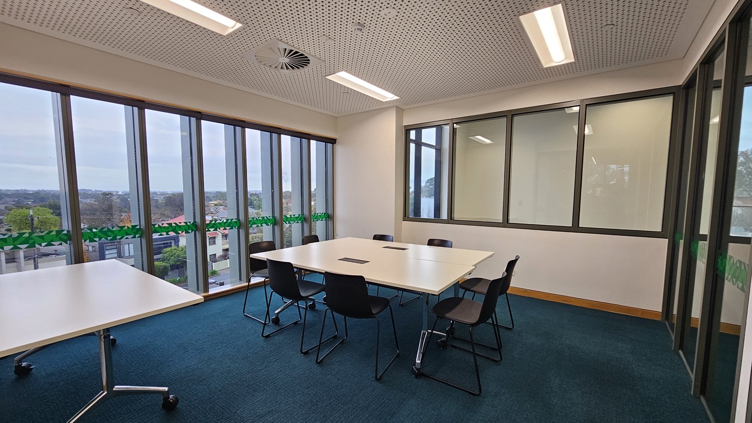 On the left, there are large windows with vertical blinds, offering a view of trees and buildings. In the middle, there is a square white table with black chairs around it. On the right, there is a wall with large glass panels and a door, providing a view into an adjacent room. The floor is covered in dark blue carpet, and the ceiling has white perforated panels with recessed lighting and a circular air vent.