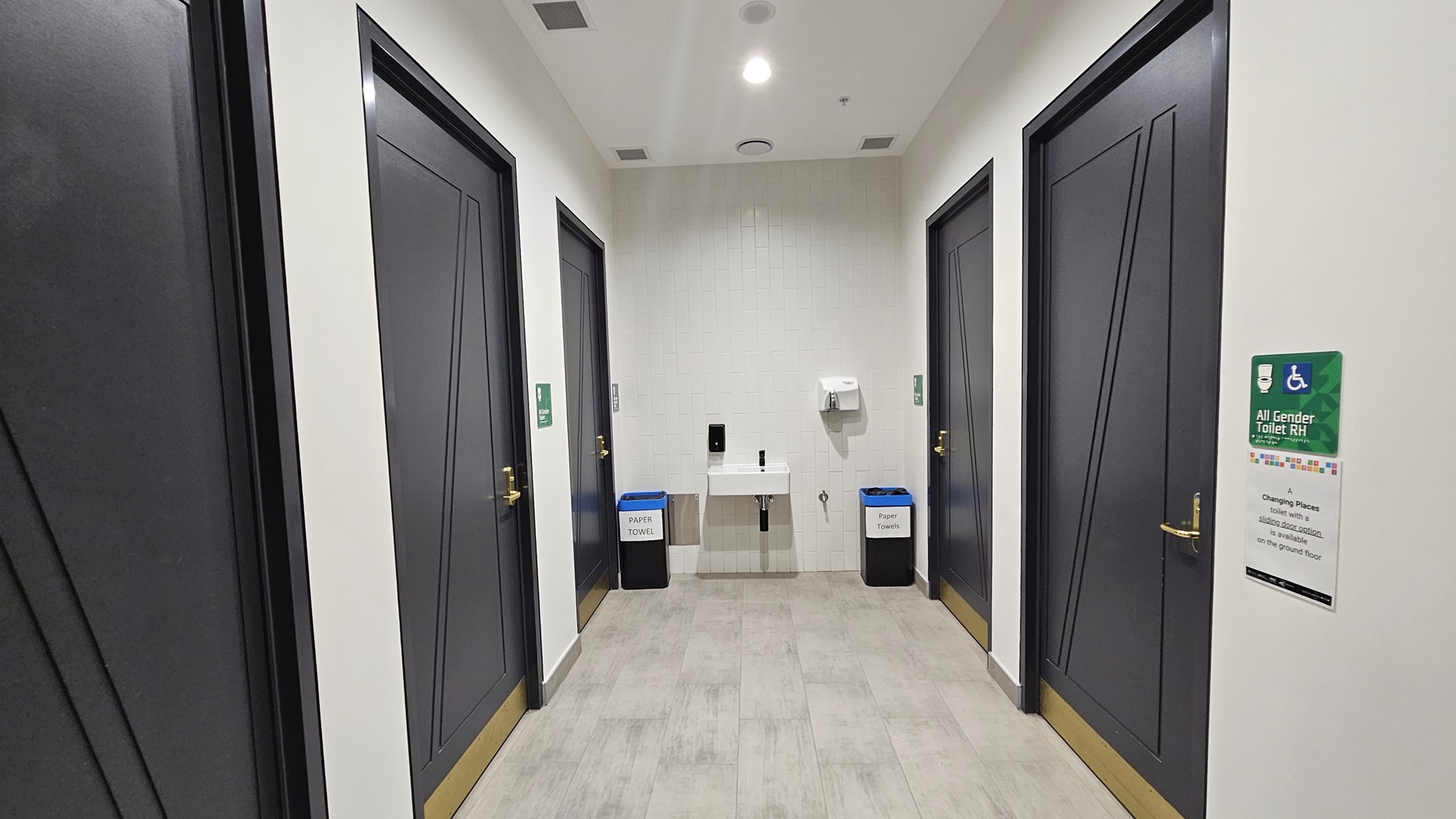 On the left, there are two dark grey doors with gold handles and green signs. In the middle, there is a white sink with a soap dispenser and paper towel dispenser above it, flanked by two bins labelled for waste and recycling. On the right, there are two more dark grey doors with gold handles and green signs, along with a wall-mounted sign displaying information. The floor is covered in light grey tiles, and the ceiling is white with recessed lighting and a circular air vent.