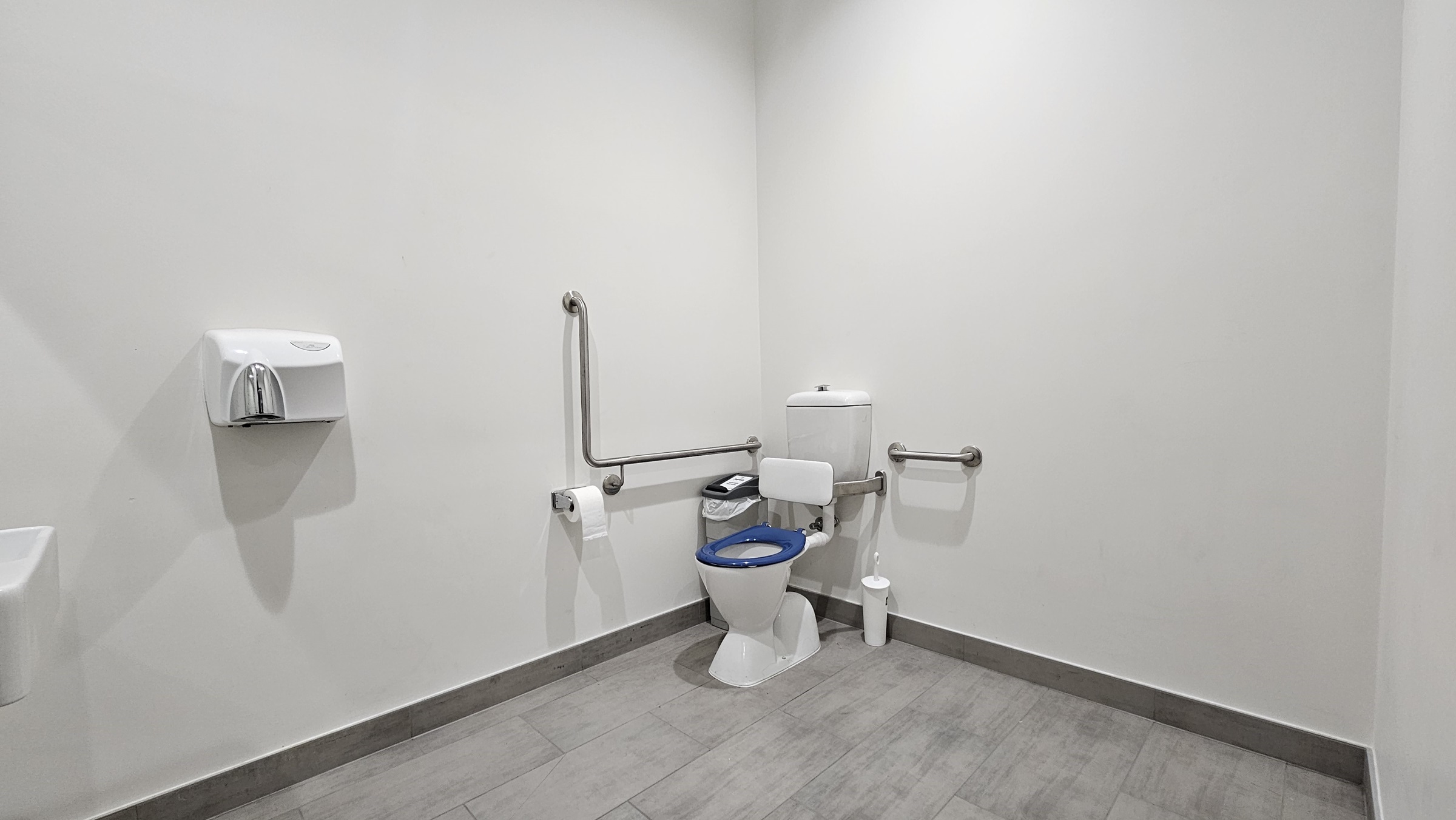On the left, there is a white wall-mounted soap dispenser. In the middle, there is a white toilet with a blue seat, equipped with stainless steel grab bars on both sides and a white toilet paper holder. On the right, there is a white wall with no fixtures. The floor is covered in light grey tiles, and the ceiling is white with recessed lighting.