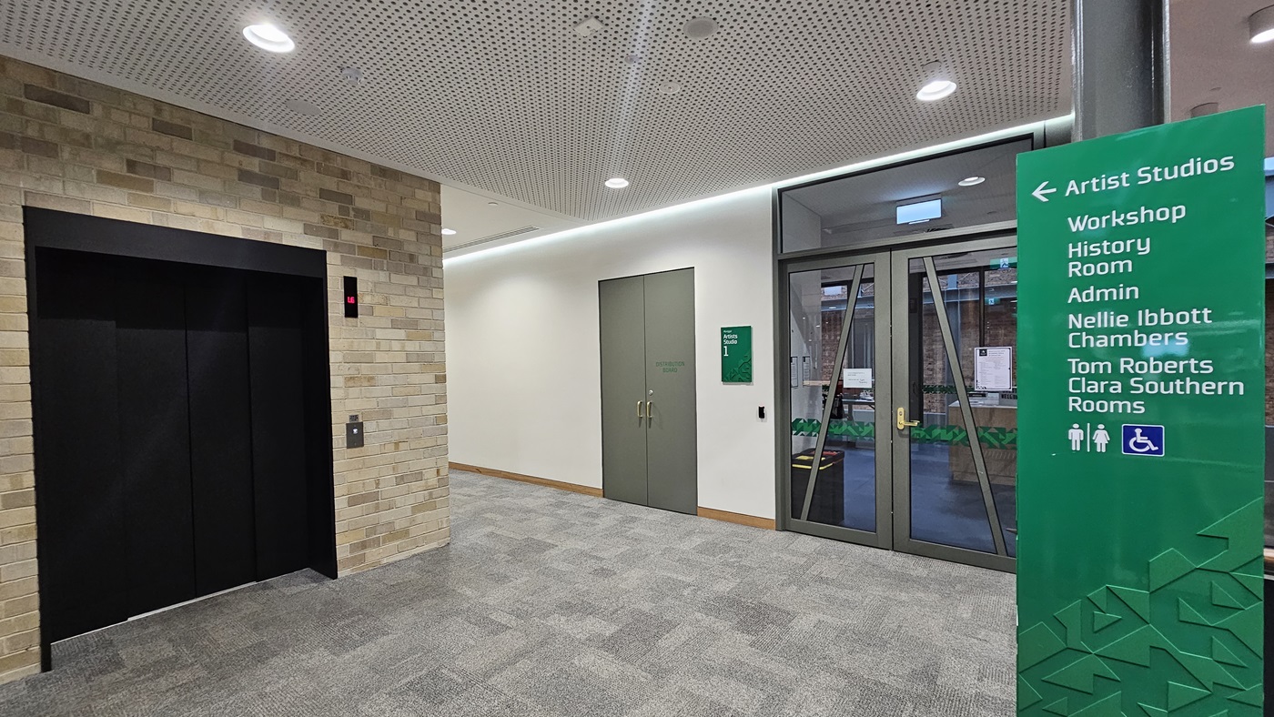 On the left, there is a black lift with a control panel and a red indicator light, set in a wall of light-coloured bricks. In the middle, there are two grey double doors with a green sign next to them. On the right, there is a glass door with a view of an outdoor area and a green sign listing room names and directions. The floor is covered in grey carpet tiles, and the ceiling is white with perforated panels and recessed lighting.