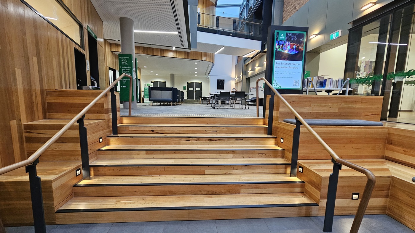 On the left, there are wooden walls with vertical panels and a green sign indicating directions. In the middle, there is a wide wooden staircase with black handrails and integrated lighting on the steps, leading to an open area with seating and tables. On the right, there is a digital information screen and more wooden panelling. The floor is covered with grey tiles, and the ceiling is white with recessed lighting and wooden accents.
