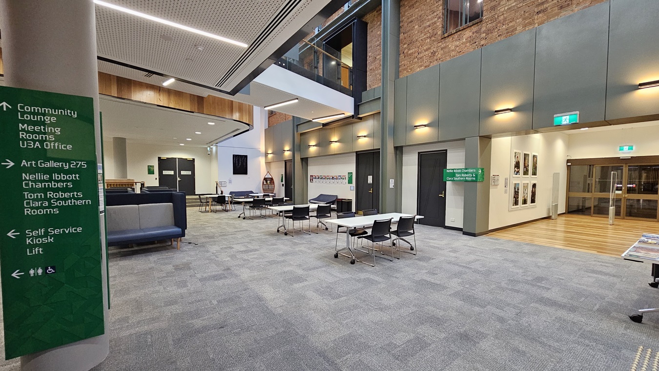 On the left, there is a green sign listing various rooms and facilities. In the middle, there are tables and chairs arranged in an open lounge area with a grey carpeted floor. In the background, there are black double doors and a staircase leading to an upper level with glass railings. On the right, there is a brick wall with framed pictures, a green exit sign, and a wooden reception counter with brochures. The ceiling is white with recessed lighting and a perforated panel design.