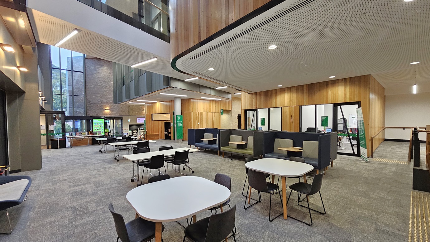 On the left, there are tall windows and a seating area with benches and tables. In the middle, there are several tables and chairs arranged in an open lounge area with grey carpeted flooring. In the background, there is a reception desk with a green sign and a brick wall. On the right, there are enclosed office spaces with glass walls and wooden panelling. The ceiling is white with recessed lighting and perforated panels.