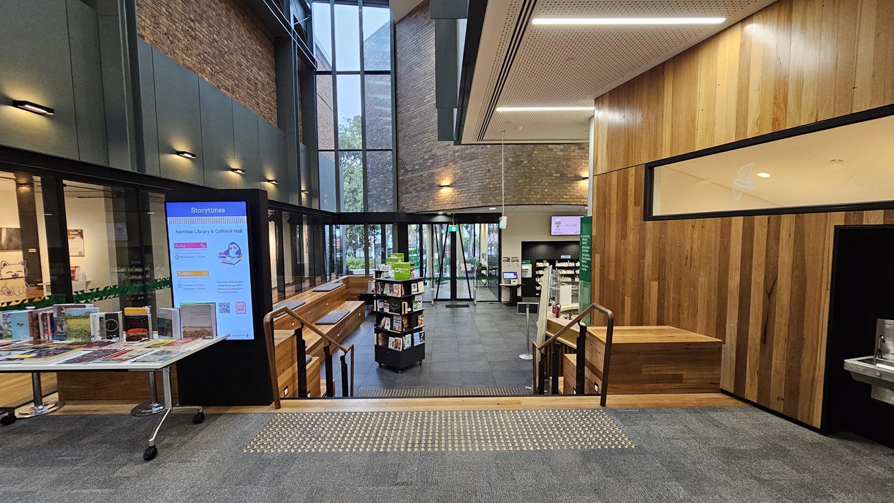 On the left, there is a tall digital sign displaying information next to a table with books and brochures. In the middle, there is a staircase with wooden handrails leading down to a lower level with bookshelves and a seating area. In the background, there are large windows and a glass door. On the right, there is a wooden wall with a window into an office space and a water fountain. The floor is a mix of grey tiles and grey carpet, and the ceiling has recessed lighting and perforated panels.
