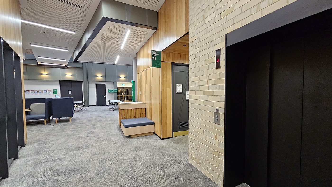 On the left, there are seating areas with blue upholstered chairs and a long table with chairs. In the middle, there is a wooden reception desk with a green sign above it and a bench with grey cushions. In the background, there are more seating areas and a green exit sign. On the right, there is a brick wall with a black lift door. The floor is covered with grey carpet tiles, and the ceiling has recessed lighting and perforated panels.