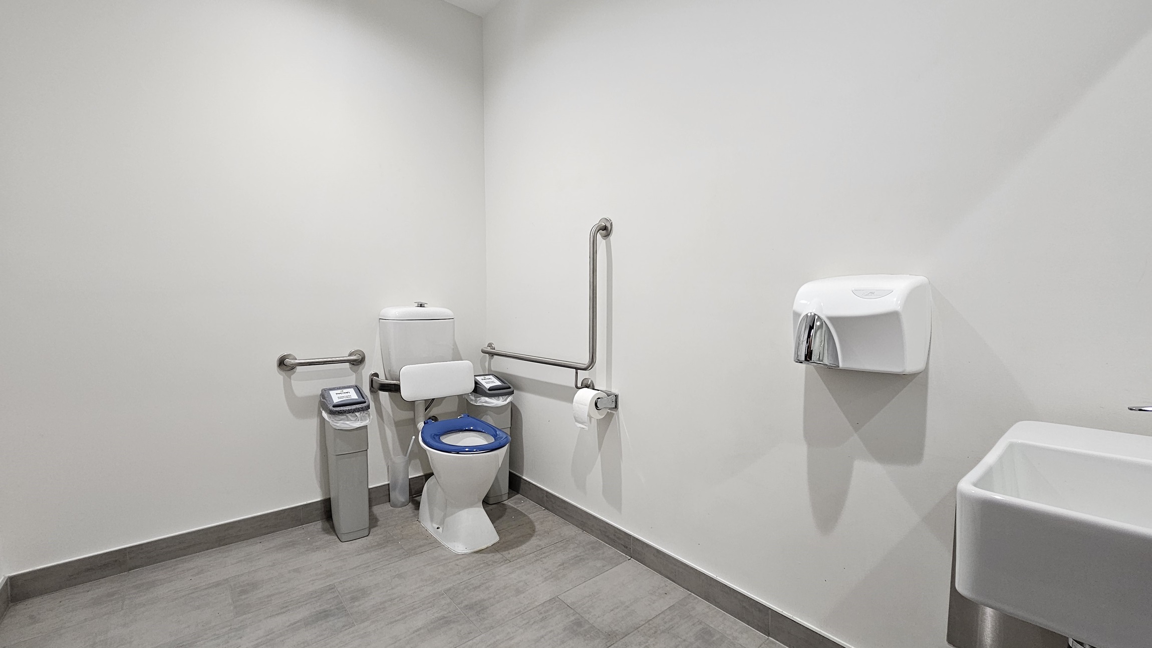 On the left, there is a white toilet with a blue seat, flanked by two grey bins and metal grab bars. In the middle, there is a toilet paper holder attached to the wall. On the right, there is a white wall-mounted paper towel dispenser above a white sink. The floor is covered with grey tiles, and the ceiling is white with recessed lighting.