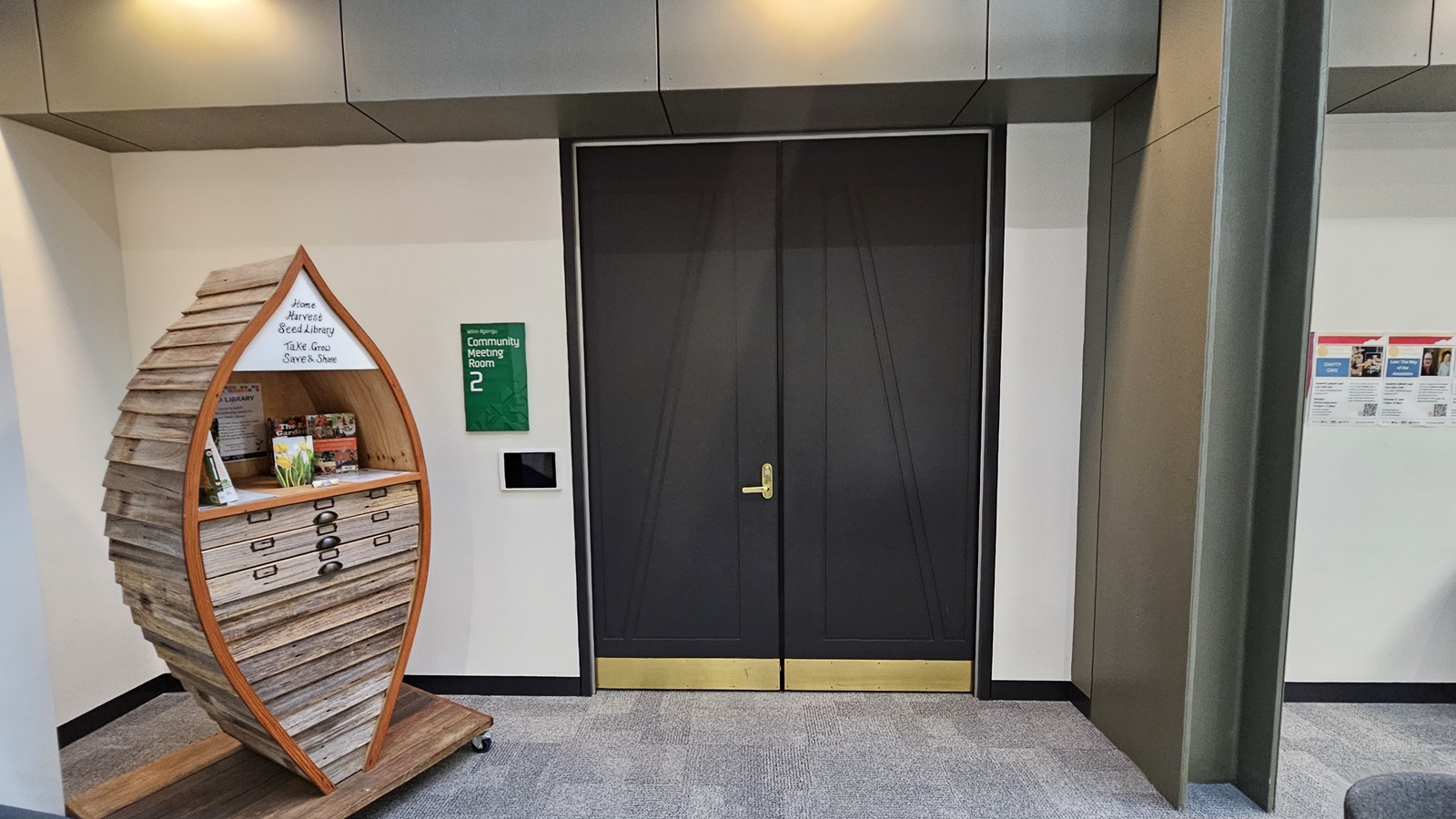 On the left, there is a wooden display shaped like a boat, containing brochures and drawers, with a sign on top. In the middle, there are large black double doors with gold handles and a green sign to the right of the doors. On the right, there is a white wall with a bulletin board displaying various notices. The floor is covered with grey carpet tiles, and the ceiling has recessed lighting.