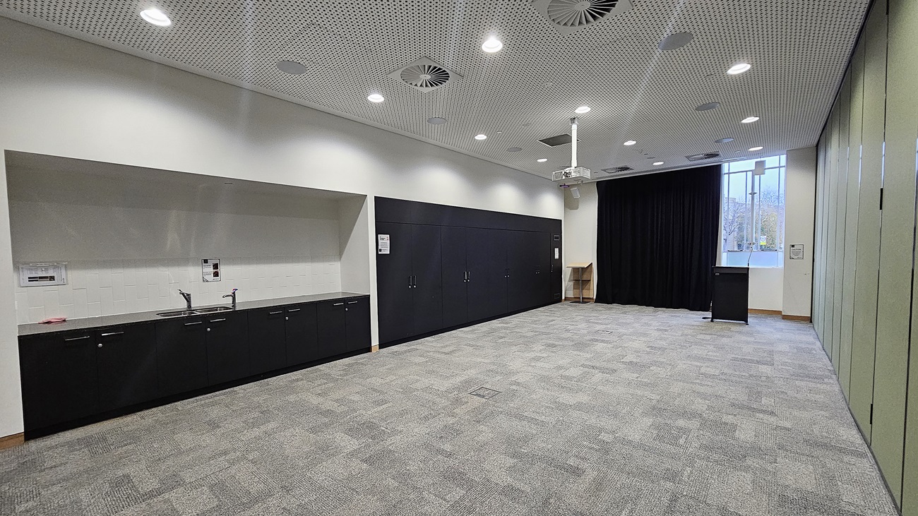 On the left, there is a long black counter with a sink, cabinets, and a white backsplash. In the middle, there is an open space with grey carpet tiles. In the background, there is a black curtain, a podium, and a window with a view of trees. On the right, there is a green partition wall. The ceiling is white with recessed lighting, circular air vents, and perforated panels.