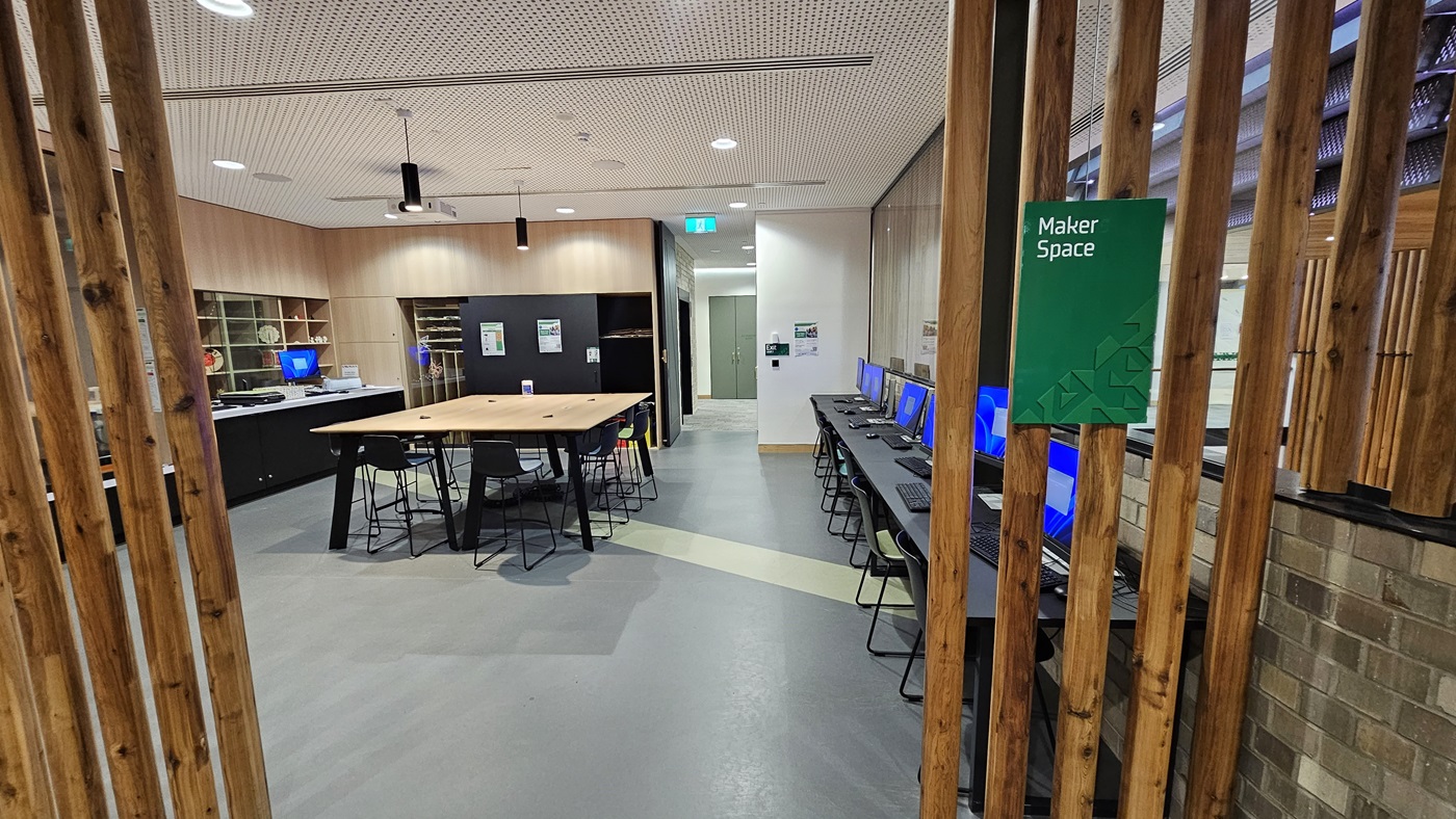 On the left, there is a white wall with a green exit sign above a door. In the middle, there is a long, L-shaped bench with blue and green cushions, and a round wooden table in front of it. On the right, there is a counter with a black top and light wood finish. The floor is covered in grey carpet tiles, and the ceiling has white perforated panels with recessed lighting and black cylindrical lights.