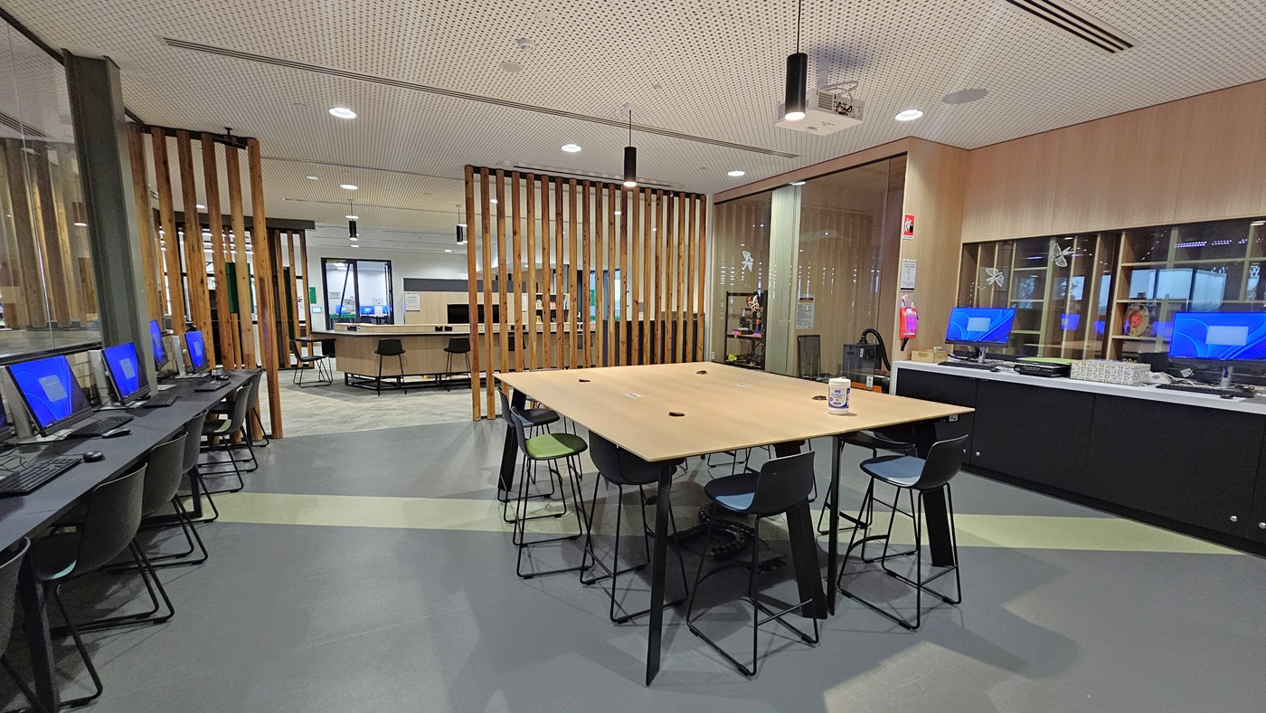 On the left, there are computer workstations with black chairs and blue screens, next to a wall with vertical wooden slats. In the middle, there is a large wooden table with black and green stools around it. On the right, there is a black counter with computers and a sink, backed by light wood cabinets. The floor is grey with green accents, and the ceiling has white perforated panels with recessed lighting and black cylindrical lights.