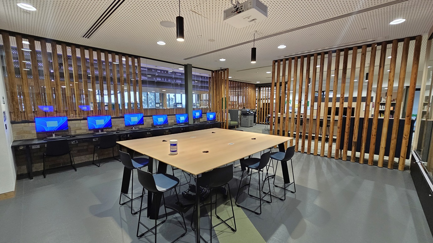 On the left, there are computer workstations with blue screens and black chairs, positioned against a wall with vertical wooden slats. In the middle, there is a large wooden table with black stools around it and a container in the centre. On the right, there is a continuation of the vertical wooden slats and a glimpse of more workstations. The floor is grey with green accents, and the ceiling has white perforated panels with recessed lighting and a projector.