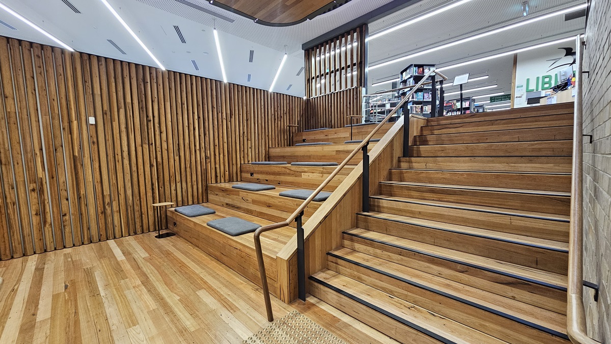 On the left, there is a lift with a "Staff Only" sign next to it, a grey column, and a long table with black chairs near large windows. In the middle, there is a staircase with wooden steps and rainbow-coloured stripes, leading to an upper level with a glass railing. On the right, there is a white wall with a door and a sign for "Workspaces." The floor is covered in grey tiles, and the ceiling is white with a perforated design and wooden panels with recessed lighting.