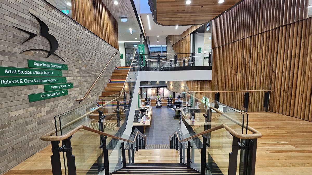 On the left, there is a brick wall with green directional signs and a black bird sculpture. In the middle, there is a staircase with wooden steps and glass railings leading down to a lower level with bookshelves and a reception desk. On the right, there is a wooden wall with vertical slats and a glass railing on the upper level. The floor is made of light-coloured wood, and the ceiling has white panels with recessed lighting and wooden sections.