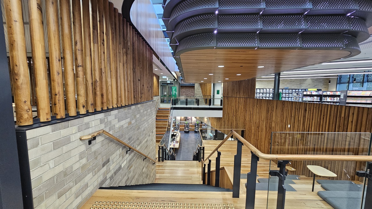 On the left, there is a staircase with wooden steps and a handrail, bordered by a wall with light grey bricks and vertical wooden slats above. In the middle, the staircase leads down to a lower level with bookshelves and tables. On the right, there is a continuation of the wooden slat wall and glass railings. The floor is wooden, and the ceiling has white panels with recessed lighting and a large, curved, perforated metal structure.