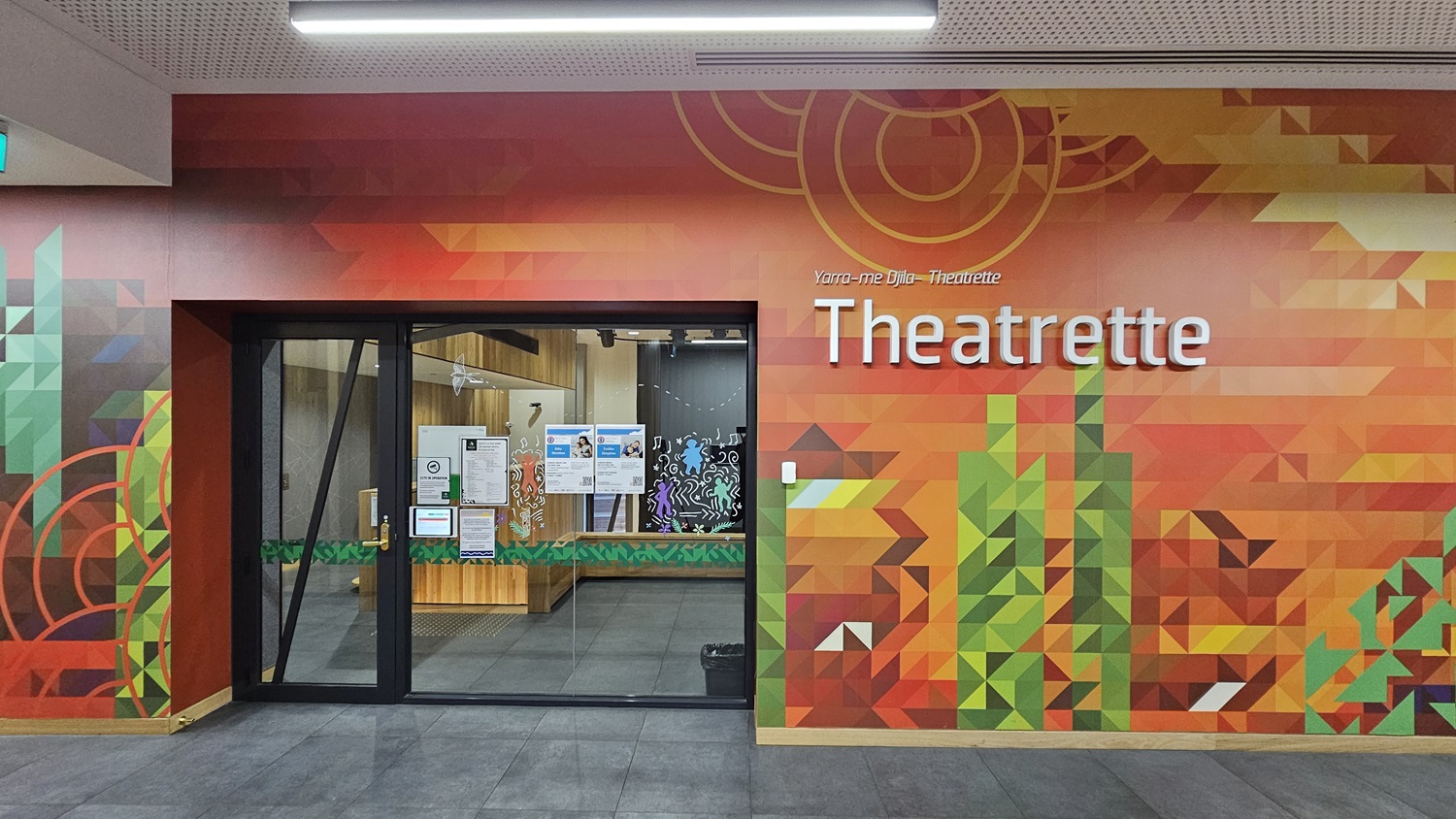 On the left, there is a curved wooden wall with vertical panels leading to an open area with desks and chairs. On the right, a colourful geometric mural in shades of orange, green, and red covers the wall, with the word "Theatrette" in large white letters. In the middle, a glass door with a black frame provides entry to the theatrette. The floor is covered with large grey tiles, and the ceiling is white with a perforated design and recessed lighting.