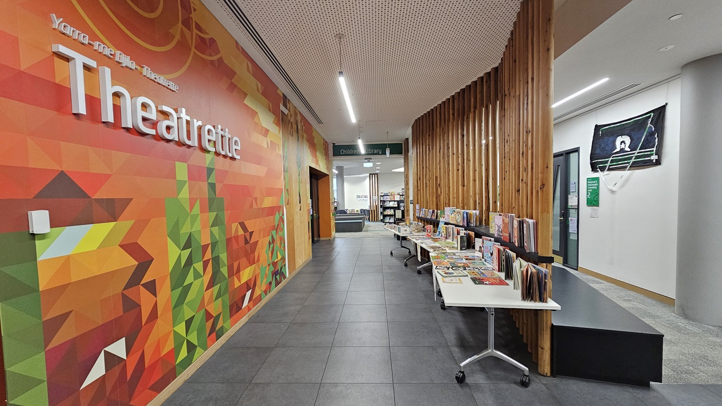 On the left, there is a vibrant wall mural with geometric patterns in green, orange, and red, featuring the word "Theatrette" in large white letters. On the right, there are wooden slats forming a partial partition, with tables displaying various books and magazines. In the middle, a hallway leads to a room with bookshelves. The floor is covered with large grey tiles, and the ceiling is white with a perforated design and linear lighting fixtures.