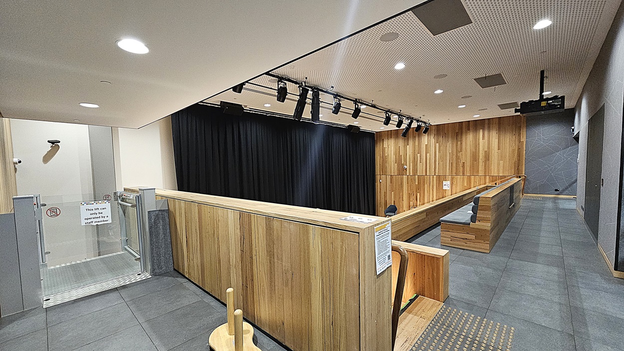 On the left, there is a glass door with a sign and a small wooden stool nearby. On the right, a wooden-panelled wall extends along the length of the room, with grey tiles on the lower part. In the middle, there is a stage area with black curtains and overhead stage lighting. The floor is covered with large grey tiles, and the ceiling is white with a perforated design and recessed lighting.