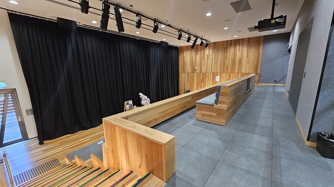 On the left, there is a wooden staircase with colourful stripes on the steps, leading up to a stage area with black curtains and overhead stage lighting. On the right, a wooden-panelled wall extends along the length of the room, with grey tiles on the lower part. In the middle, there is a long wooden bench with a few items placed on it. The floor is covered with large grey tiles, and the ceiling is white with a perforated design and recessed lighting.