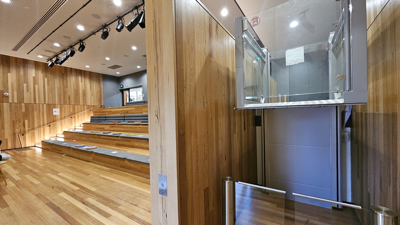 On the left, there are tiered wooden seating steps with grey cushions, leading up to a stage area with black stage lighting fixtures hanging from the ceiling. On the right, there is a glass-enclosed lift with metal railings. In the middle, a wooden wall separates the seating area from the lift. The floor is made of polished wooden planks, and the ceiling is white with a perforated design and recessed lighting.