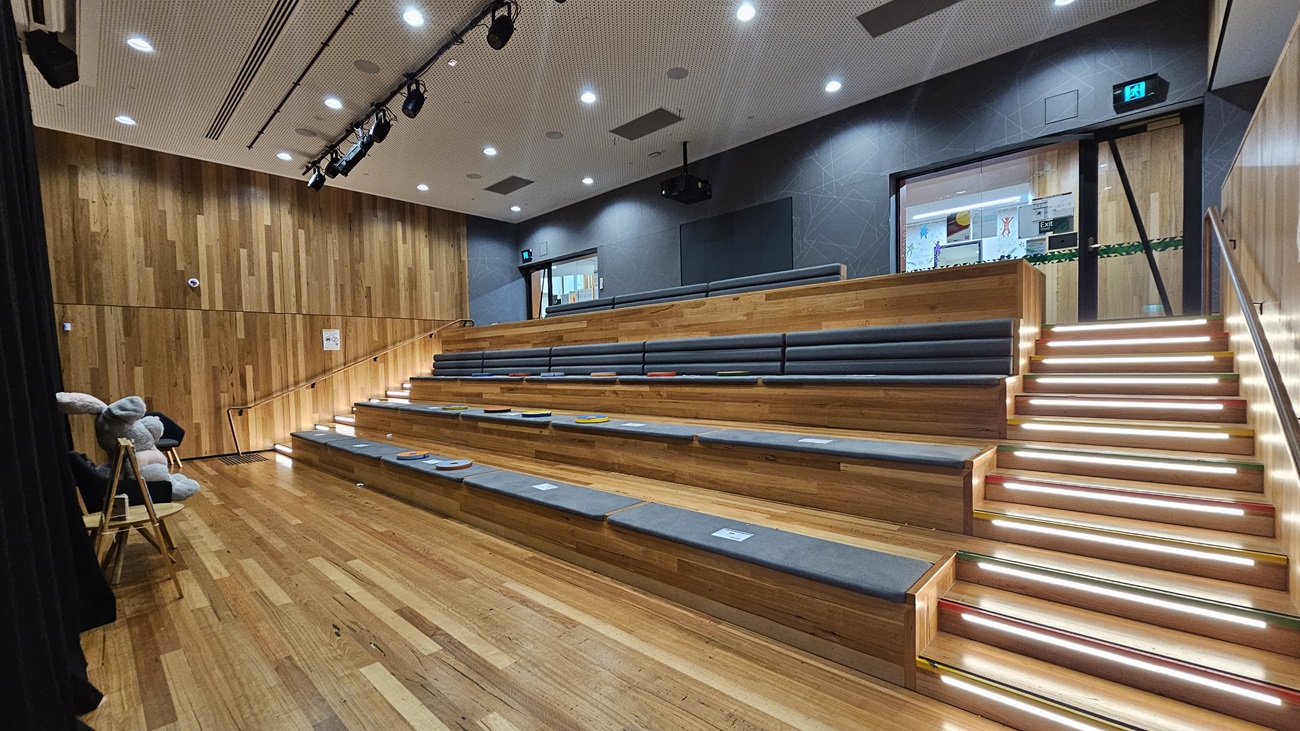 On the left, there are tiered wooden seating steps with grey cushions, leading up to a stage area with black stage lighting fixtures hanging from the ceiling. On the right, a staircase with illuminated steps and a glass railing ascends to an upper level. In the middle, the seating area continues with additional steps and cushions. The floor is made of polished wooden planks, and the ceiling is white with a perforated design and recessed lighting.