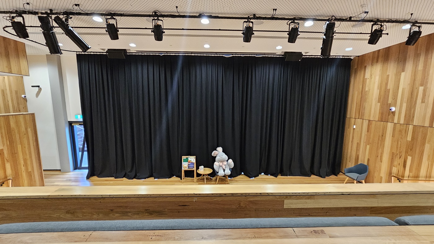 On the left, there is a wooden wall with vertical panels and a small exit door with a green emergency exit sign above it. On the right, another wooden wall with vertical panels extends to the ceiling. In the middle, there is a stage with black curtains, a small wooden table, a chair, and a large stuffed animal. The foreground features tiered wooden seating steps with grey cushions. The floor is wooden, and the ceiling is white with a perforated design and black stage lighting fixtures.