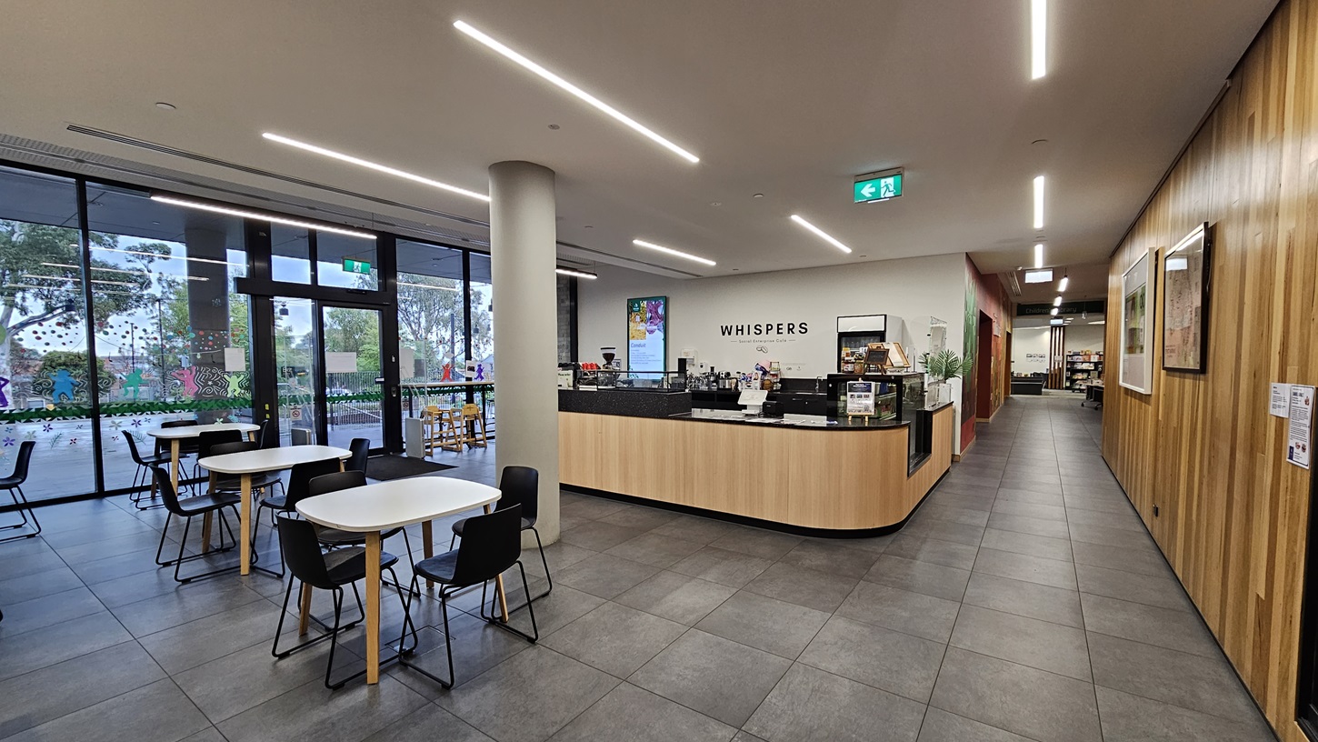 On the left, there is a lift with a "Lift Out of Order" sign and a brick wall. In the middle, there are high tables with black stools and a glass wall behind them, providing a view of the outside. On the right, a wide staircase with wooden steps and rainbow-coloured stripes on the risers, leading to the upper floor with a glass railing. The floor is covered with large grey tiles, and the ceiling is white with perforated panels and recessed lighting.