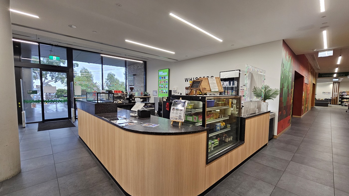 On the left, there is a large glass wall with doors leading outside, next to a cylindrical column. In the middle, there is a wooden counter with a black countertop, displaying various items and a coffee machine, with the sign "Whispers" above it. On the right, a corridor with colourful wall art and wooden panelling, leading to more rooms. The floor is covered with large grey tiles, and the ceiling is white with linear recessed lighting.