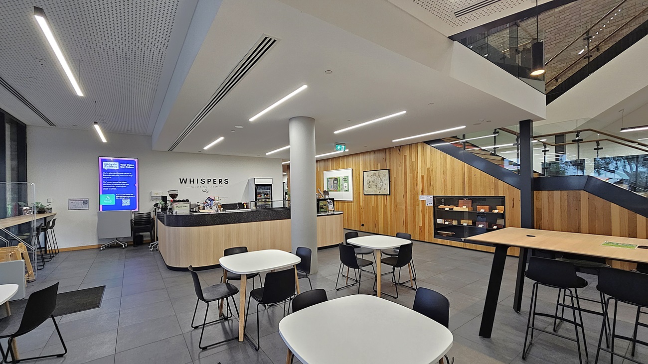 On the left, there is a digital information screen and a small counter with a coffee machine. In the middle, there is a cylindrical column and a wooden counter with the sign "Whispers" above it, surrounded by white tables and black chairs. On the right, there is a staircase with wooden steps and a glass railing leading to the upper floor, with wooden panelling on the wall. The floor is covered with large grey tiles, and the ceiling is white with linear recessed lighting and perforated panels.