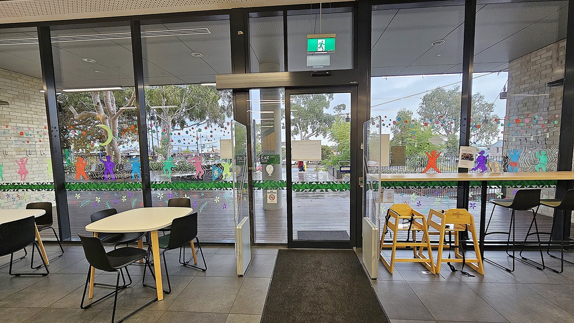 On the left, there are round white tables with black chairs. In the middle, there is a glass door with a green exit sign above it, flanked by large glass windows decorated with colourful paper figures and garlands. On the right, there is a wooden counter with high chairs facing the window. The floor is covered with large grey tiles, and the ceiling is white with perforated panels.