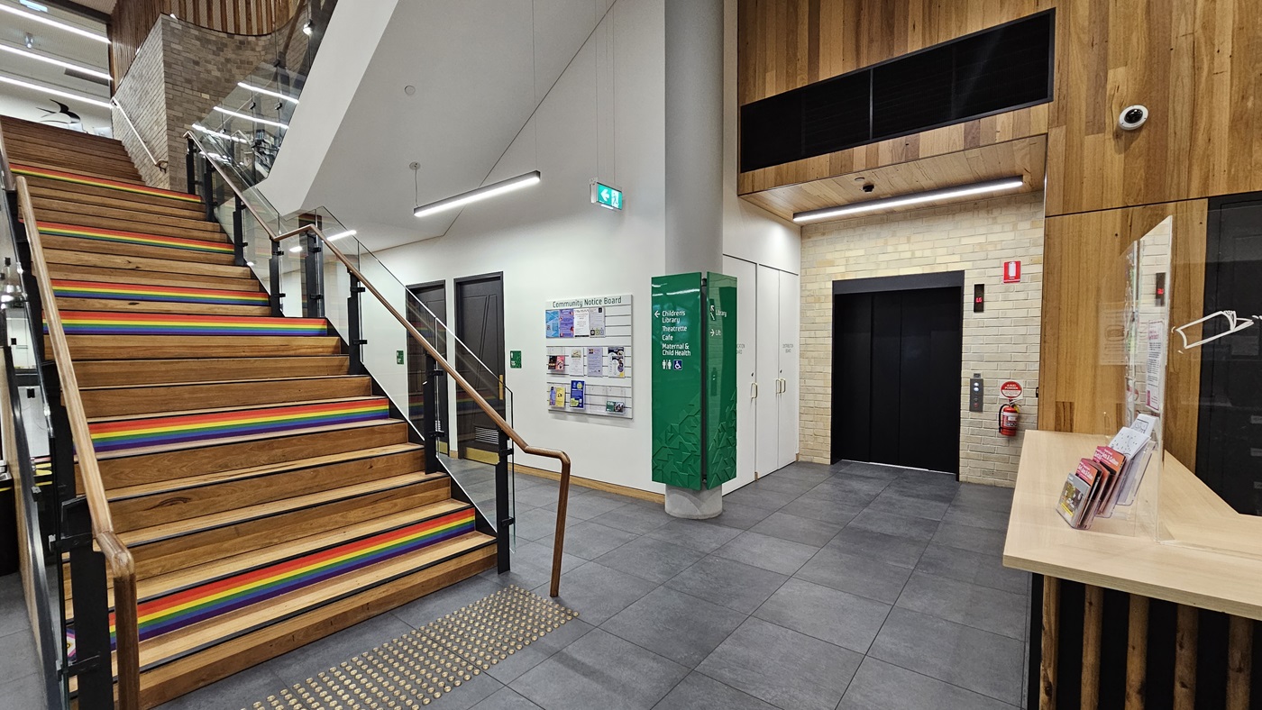 On the left, there is a staircase with wooden steps and rainbow-coloured stripes on the risers, leading to the upper floor with a glass railing. In the middle, there is a cylindrical column and a green information board on the wall. On the right, there is a lift with a black door, a fire extinguisher, and a wooden counter with brochures. The floor is covered with large grey tiles, and the ceiling is white with recessed lighting and wooden panelling near the lift.