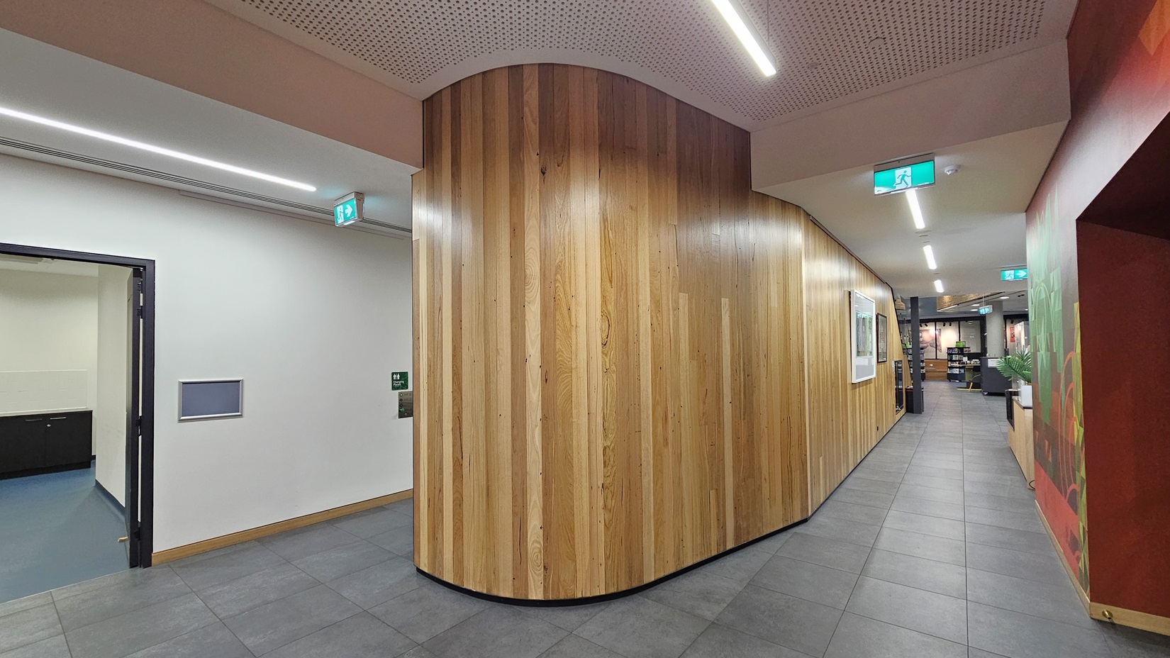 On the left, there is a doorway with a black frame leading to a room with a blue floor, and an emergency exit sign above it. In the middle, there is a curved wooden wall with vertical panels. On the right, there is a corridor with grey floor tiles, a white ceiling with perforated panels and linear recessed lighting, and another emergency exit sign. The corridor extends into the background, where more rooms and decorations are visible.