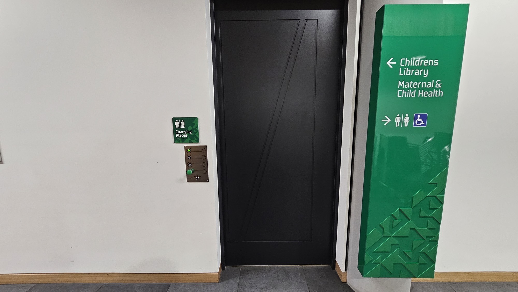 On the left, there is a white wall with a small green sign indicating "Change Project Room" and an intercom with buttons. In the middle, there is a black door with a geometric pattern. On the right, there is a large green sign with white text pointing to the "Children's Library" and "Maternal & Child Health" with icons for accessibility. The floor is covered with grey tiles, and the baseboard is wooden.